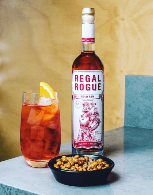 Highball glass with red cocktail over ice and orange wedge, bowl of nuts and bottle of Regal Rogue Bold Red vermouth on counter top and yellow background