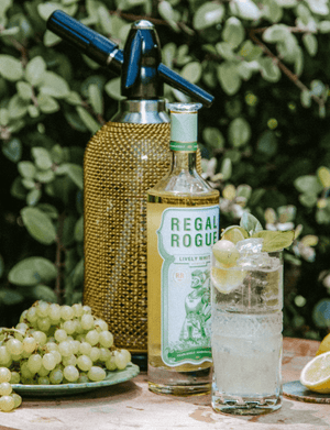 Highball glass with grapes on side plate and soda siphon and Regal Rogue Lively White bottle in the middle