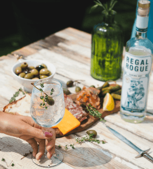 Hand holding a vermouth spritz wine glass with olives and charcuterie in the background and bottle of Regal Rogue Daring Dry vermouth