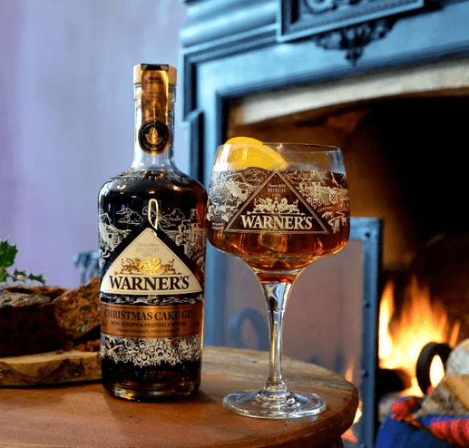 Warner's Christmas Cake Gin in glass with cola garnished with orange next to bottle in front of fire